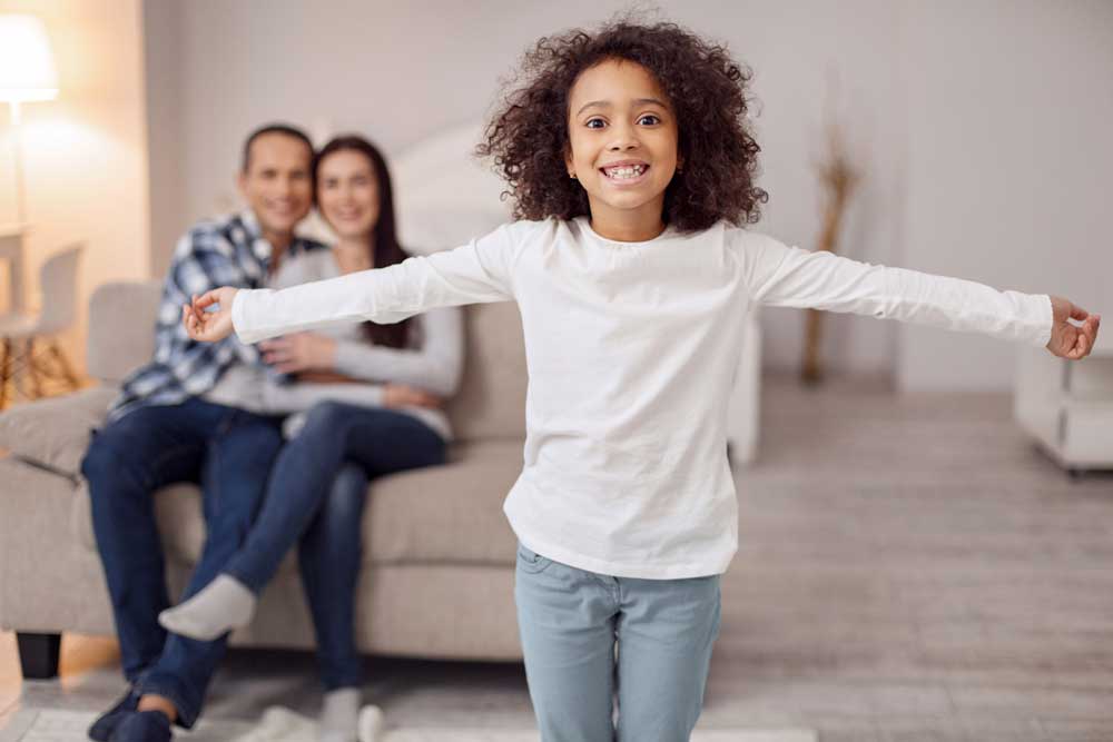 Pretty cheerful curly-haired girl smiling and spreading her arms and her parents sitting on the couch in the background